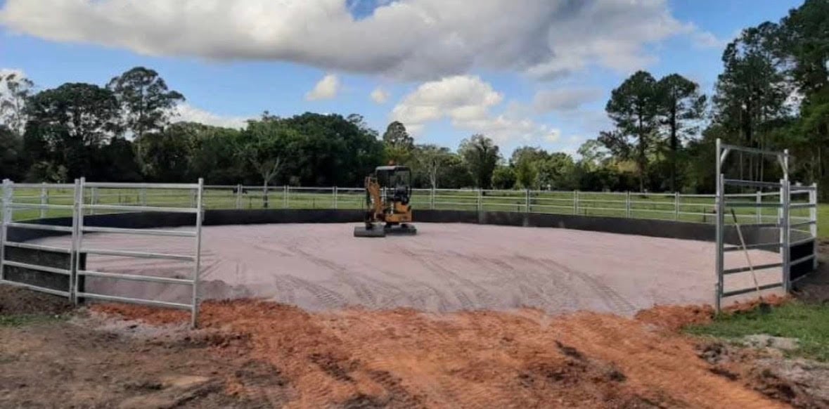 A tractor working on an arena floor inside a fenced equestrian riding ring with white gates and red dirt surrounding it.
